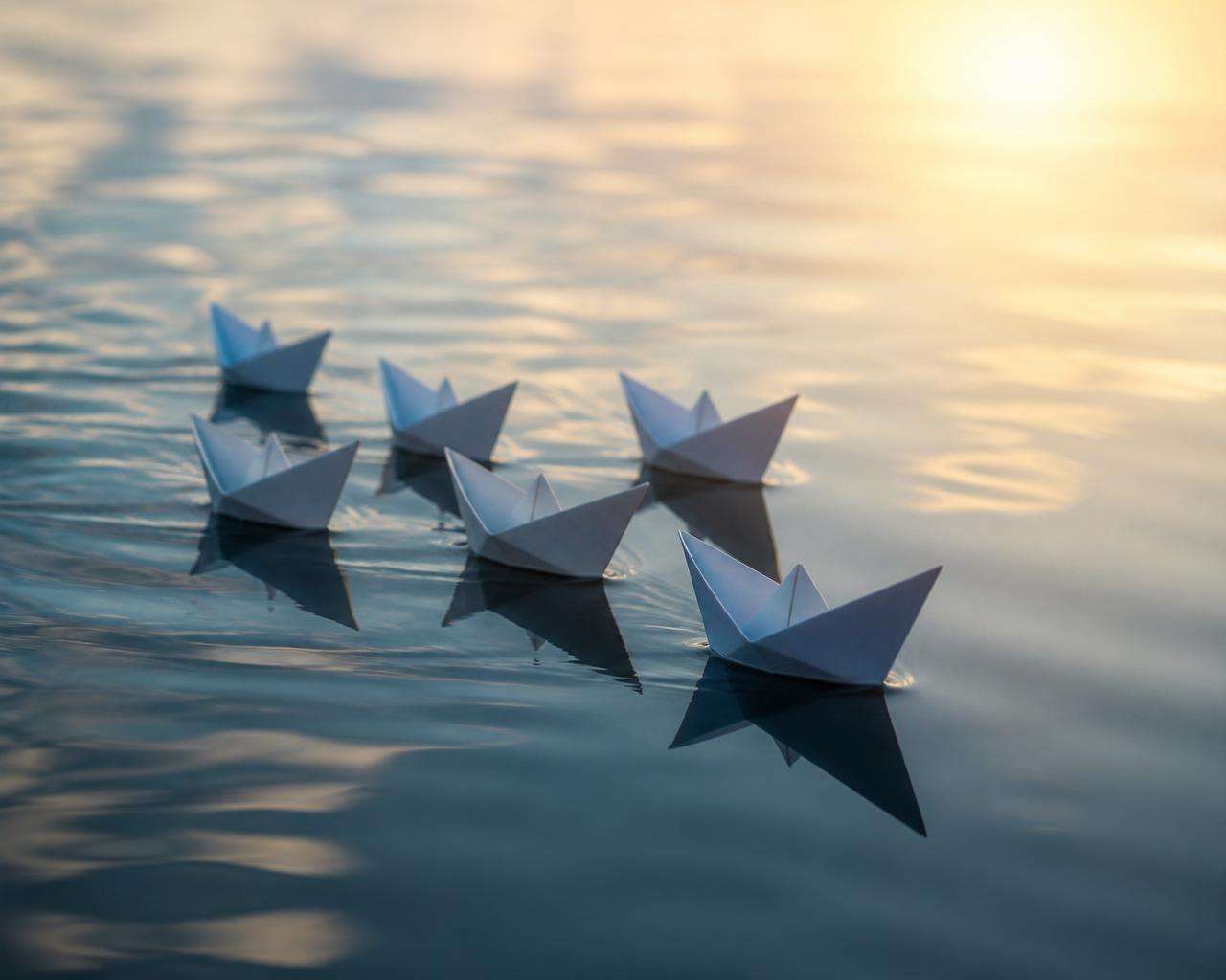 A small group of paper boats moving together in the same direction on calm reflective water — a symbol of independent people moving with a shared sense of direction
